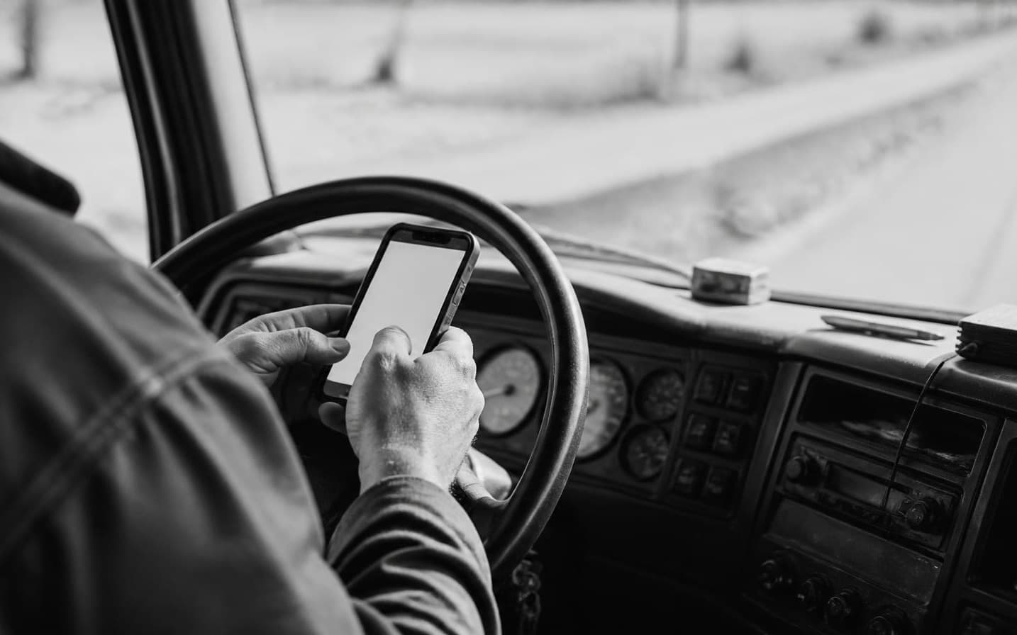 Las manos de un chofer sosteniendo un teléfono contra el volante, con el camión detenido y la ruta adelante.